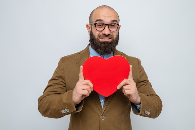 Bearded man in brown suit wearing glasses holding heart made from cardboard looking smiling cheerfully happy and positive