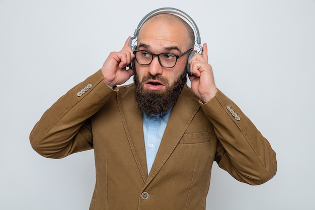 Bearded man in brown suit wearing glasses with headphones looking surprised smiling enjoying his favorite music standing over white background