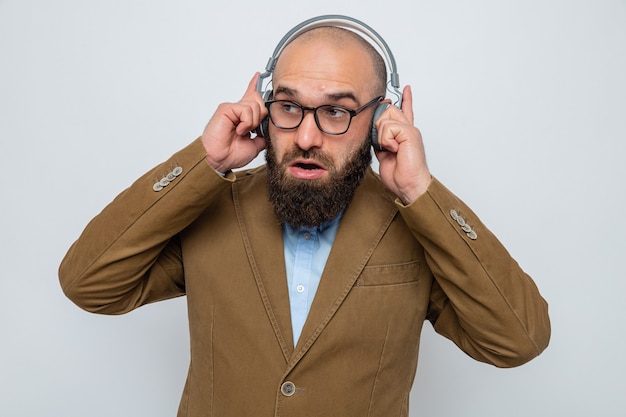 Uomo barbuto in abito marrone con gli occhiali con le cuffie che sembra sorpreso sorridente godendosi la sua musica preferita in piedi su sfondo bianco