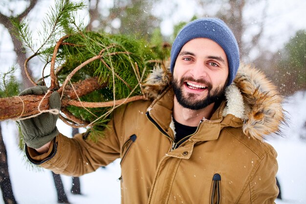 Bearded man carrying freshly cut down christmas tree in forest young lumberjack bears fir tree on