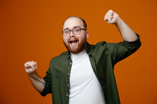 Bearded man in casual clothes clenching fists crazy happy rejoicing his success standing over orange background