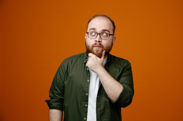 Bearded man in casual clothes looking up with pensive expression thinking hlding hand on his chin standing over orange background