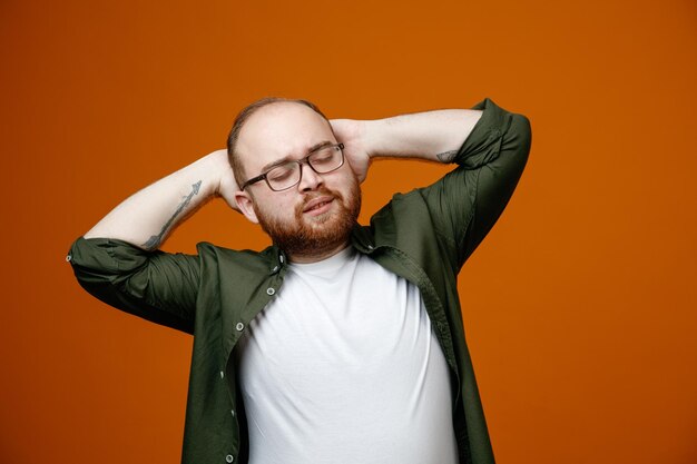 Bearded man in casual clothes wearing glasses calm and relaxed with hands behind his head standing over orange background