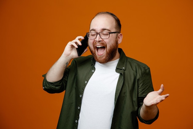 Uomo barbuto in abiti casual con occhiali felice e positivo che sorride ampiamente mentre parla al telefono cellulare in piedi su sfondo arancione