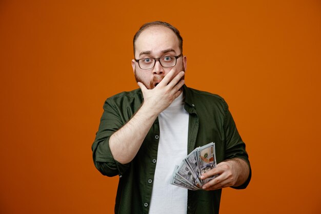 Bearded man in casual clothes wearing glasses holding cash looking at camera amazed and surprised covering mouth with hand standing over orange background