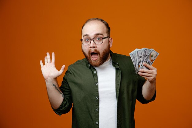 Bearded man in casual clothes wearing glasses holding cash raising arm looking at camera amazed and surprised standing over orange background