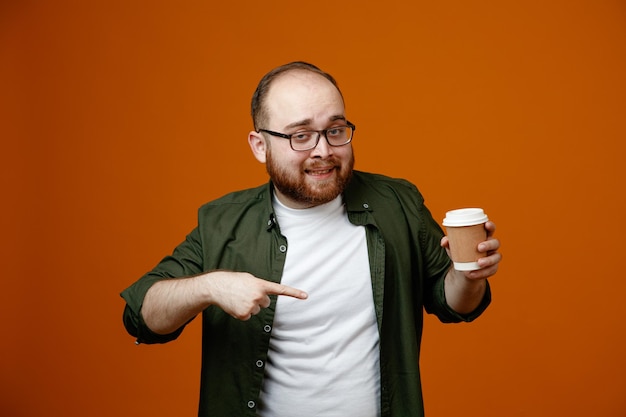 Uomo barbuto in abiti casual con gli occhiali con in mano una tazza di caffè che punta con il dito indice su di esso guardando la fotocamera sorridente felice e positivo in piedi su sfondo arancione