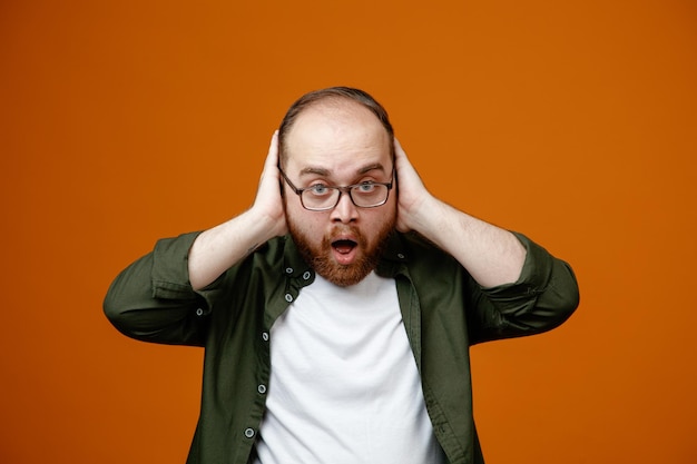 Bearded man in casual clothes wearing glasses looking at camera being confused and surprised holding hands on his head standing over orange background