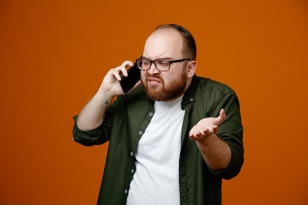 Bearded man in casual clothes wearing glasses looking dissatisfied raising arm in displeasure while talking on mobile phone standing over orange background