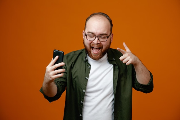 Bearded man in casual clothes wearing glasses taking selfie using smartphone showing vsign happy and cheerful standing over orange background