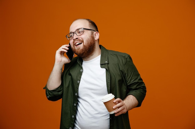 Uomo barbuto in abiti casual con gli occhiali che parla sul telefono cellulare tenendo una tazza di caffè sorridente allegramente in piedi su sfondo arancione