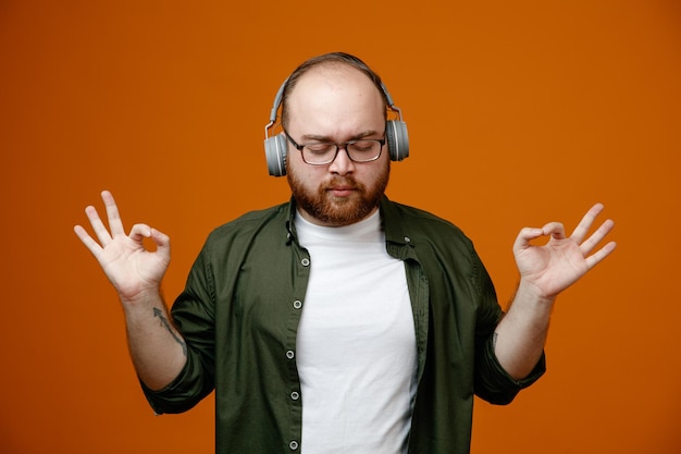 Uomo barbuto in abiti casual che indossano occhiali con le cuffie calmo e rilassato ascoltando musica facendo un gesto di meditazione con le dita in piedi su sfondo arancione