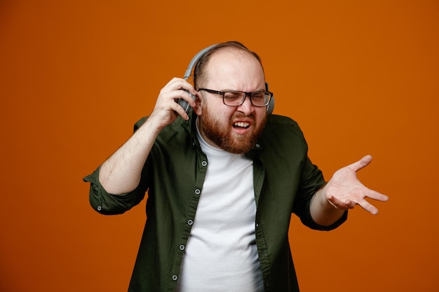 Bearded man in casual clothes wearing glasses with headphones looking confused and displeased raising arm in displeasure standing over orange background