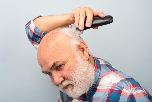 Bearded man cutting his own hair with a clipper. Man hair treatment. Gray man hair clippings. Bald man hairclipper, Mature baldness and hair loss concept.