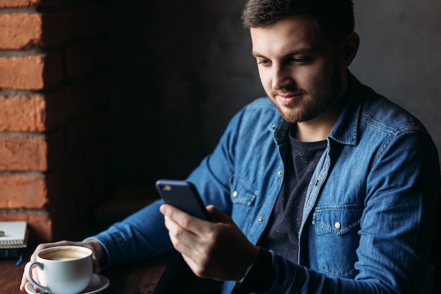 Bearded man in a denim shirt using phone inside a restaurant while drinking a coffee