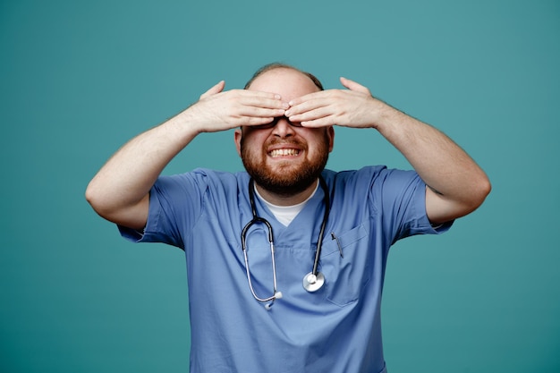 Bearded man doctor in uniform with stethoscope around neck covering eyes with hands smiling broadly standing over blue background