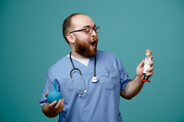 Bearded man doctor in uniform with stethoscope around neck wearing glasses holding enema and a doll looking happy and joyful standing over blue background