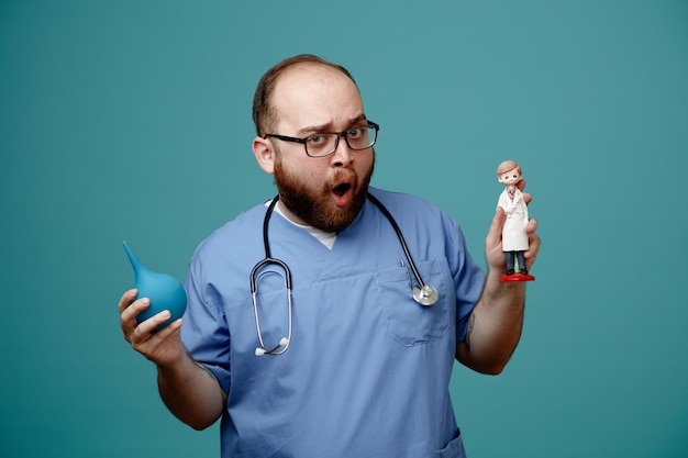 Bearded man doctor in uniform with stethoscope around neck wearing glasses holding enema and a doll looking surprised and shocked standing over blue background