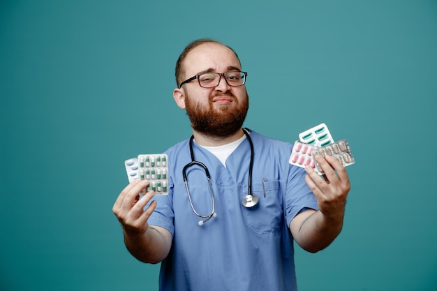 Bearded man doctor in uniform with stethoscope around neck wearing glasses holding pills looking at camera smiling confident standing over blue background