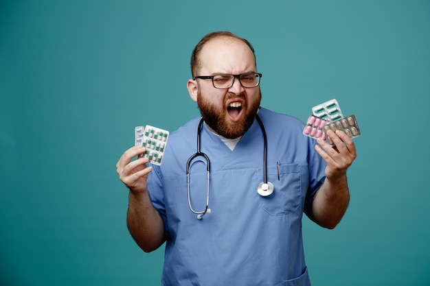 Bearded man doctor in uniform with stethoscope around neck wearing glasses holding pills shouting with aggressive expression angry and frustrated standing over blue background