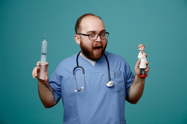 Bearded man doctor in uniform with stethoscope around neck wearing glasses holding syringe and a doll having fun smiling cheerfully standing over blue background