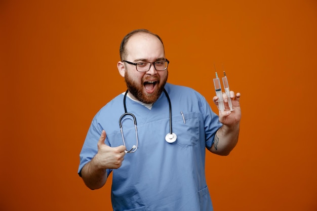 Bearded man doctor in uniform with stethoscope around neck wearing glasses holding syringes happy and excited smiling cheerfully showing thumb up standing over orange background