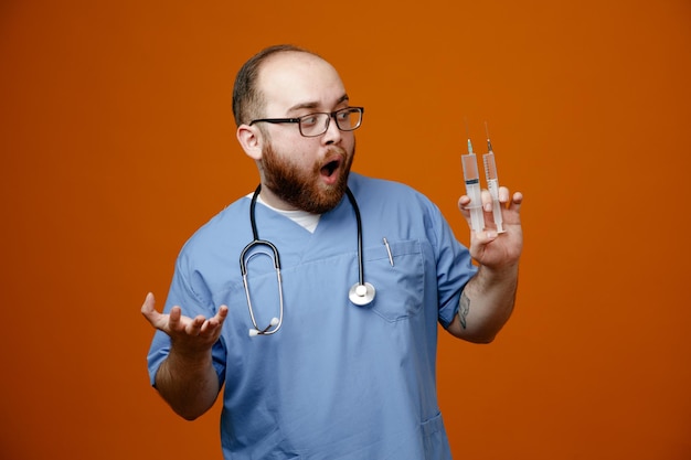 Bearded man doctor in uniform with stethoscope around neck wearing glasses holding syringes looking amazed and surprised standing over orange background