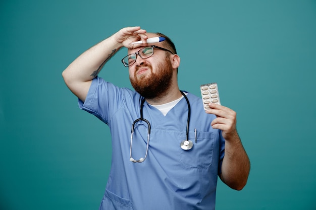 Medico uomo barbuto in uniforme con stetoscopio intorno al collo che indossa occhiali con termometro e pillole che sembrano confusi e delusi in piedi su sfondo blu