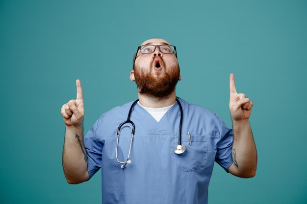 Bearded man doctor in uniform with stethoscope around neck wearing glasses looking up being surprised pointing with index fingers up standing over blue background