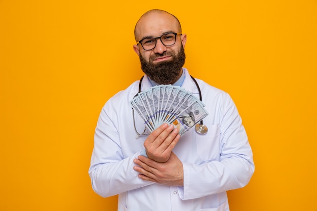 Uomo barbuto medico in camice bianco con stetoscopio intorno al collo con gli occhiali che tengono contanti guardando la telecamera sorridendo felice e contento in piedi su sfondo arancione