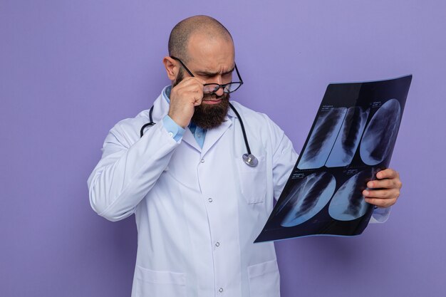 Bearded man doctor in white coat with stethoscope around neck wearing glasses holding x-ray looking at it closely concentrating