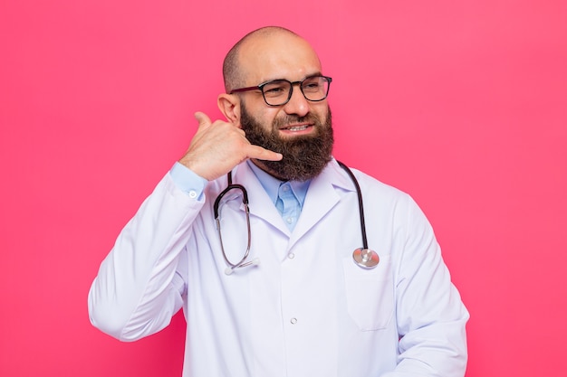 Bearded man doctor in white coat with stethoscope around neck wearing glasses looking aside smiling cheerfully making call me gesture