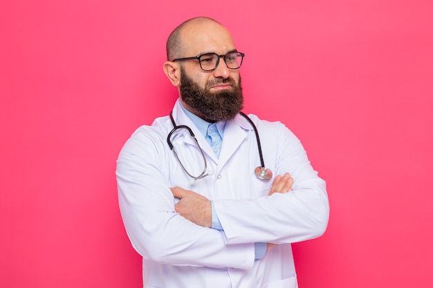 Bearded man doctor in white coat with stethoscope around neck wearing glasses looking aside with confident expression with arms crossed