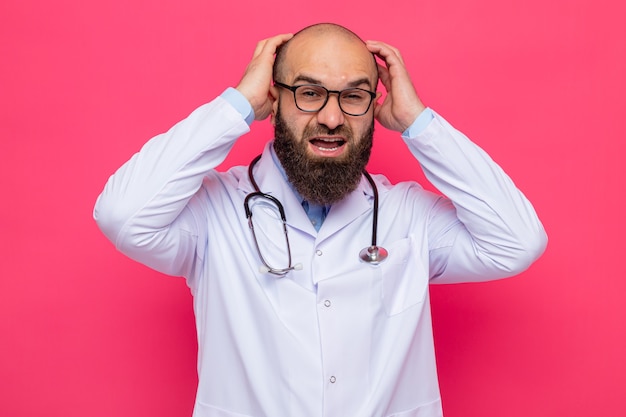 Bearded man doctor in white coat with stethoscope around neck wearing glasses looking at camera yelling with hands on his head angry and frustrated standing over pink background