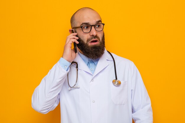 Bearded man doctor in white coat with stethoscope around neck wearing glasses looking confused while talking on mobile phone standing over orange background