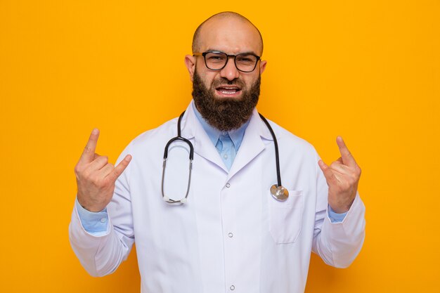 Bearded man doctor in white coat with stethoscope around neck wearing glasses looking happy and excited showing rock symbol