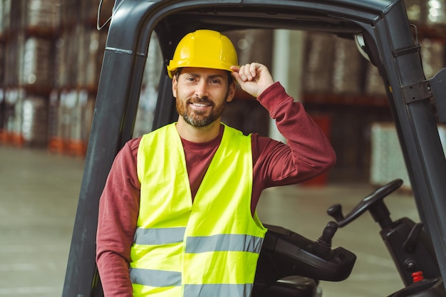 Photo bearded man driver wearing helmet standing near forklift looking at camera standing in warehouse