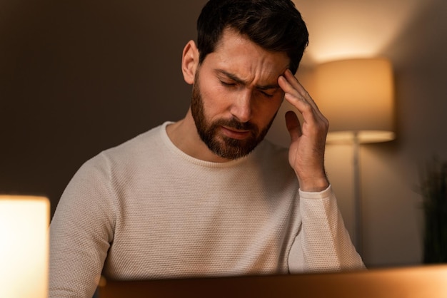 Bearded man feeling tired while sitting in front of laptop and rubbing his head during working at the evening