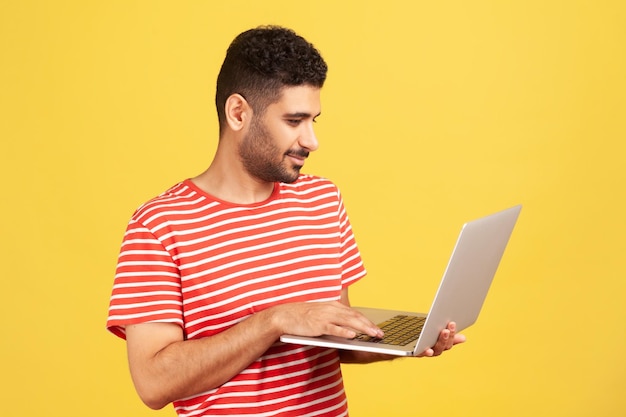 Bearded man freelancer in red striped t-shirt standing holding laptop, typing on keyboard, doing his job, teleworking on wireless gadget. Indoor studio shot isolated on yellow background