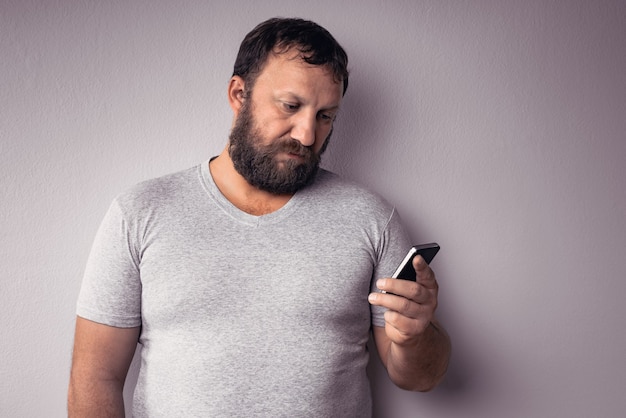 Бородатый мужчина в серой футболке держит мобильный телефон, стоя у серой стены