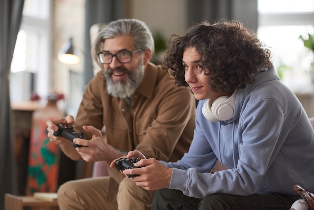 Bearded man and guy using joysticks for playing in video game while sitting on sofa at home