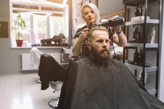 Bearded man at hairdresser with hair dryer while sitting in chair at barbershop.