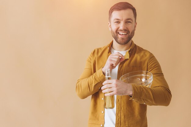 Bearded man happy and smiling in yellow shirt drinking beer and eating chips on beige background