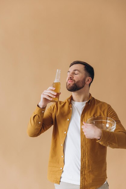 Bearded man happy and smiling in yellow shirt drinking beer and eating chips on beige background