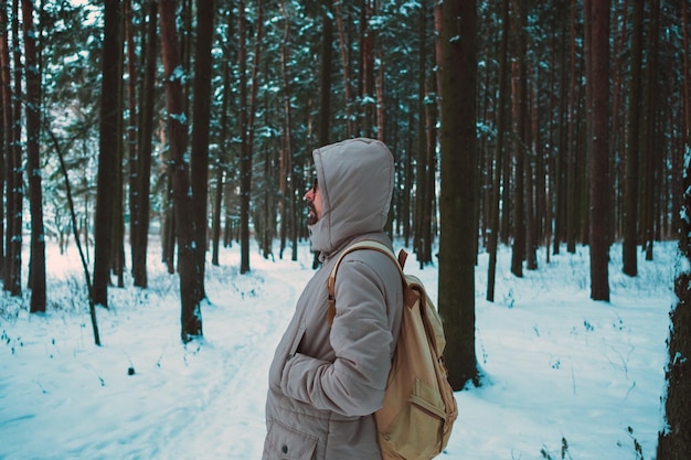 A bearded man in a hat and a backpack stands with his back in winter in a coniferous forest on the river The concept of winter travel and walks in the woods