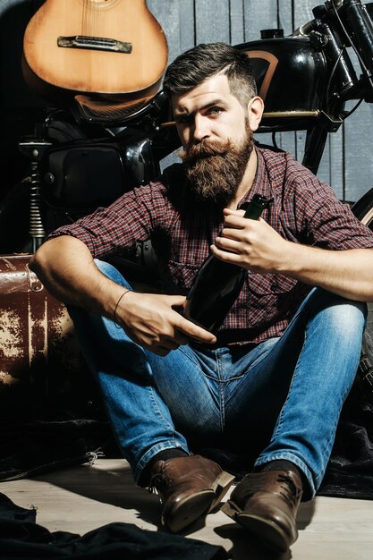 Bearded man hipster biker brutal male with beard and moustache\
in shirt sits on floor near motorcycle with wine bottle suitcase\
and guitar on wooden background