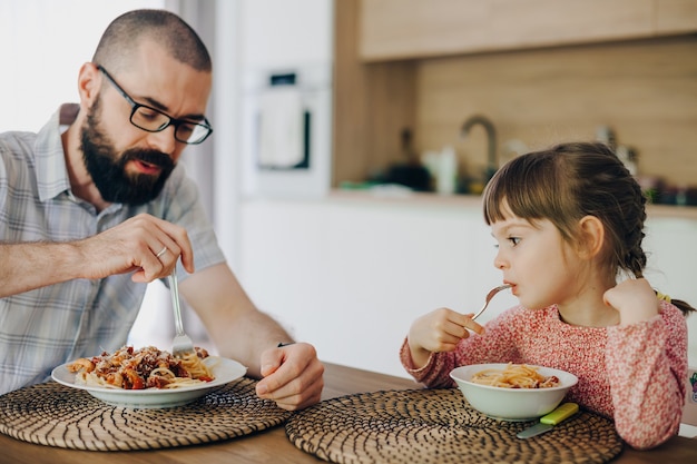 Bearded man and his cute little daughter eating spaghetti with sauce sitting in a kitchen at home