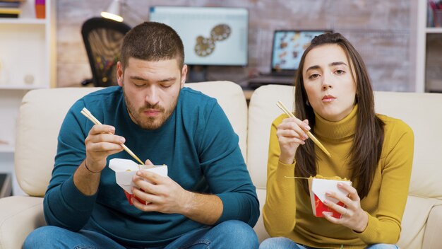 テレビを見ながら箸で麺を食べるひげを生やした男と彼のガールフレンド。