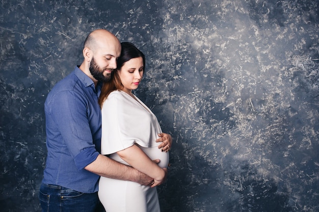 Bearded man holding his pregnant wife by belly on abstract surface.