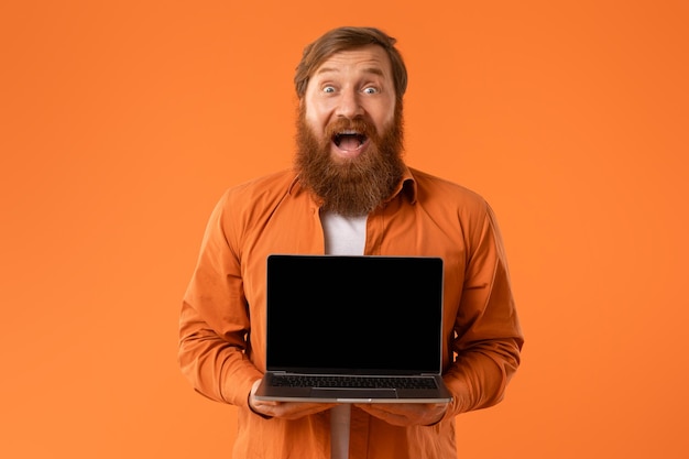 Bearded man holding laptop computer with empty screen orange background